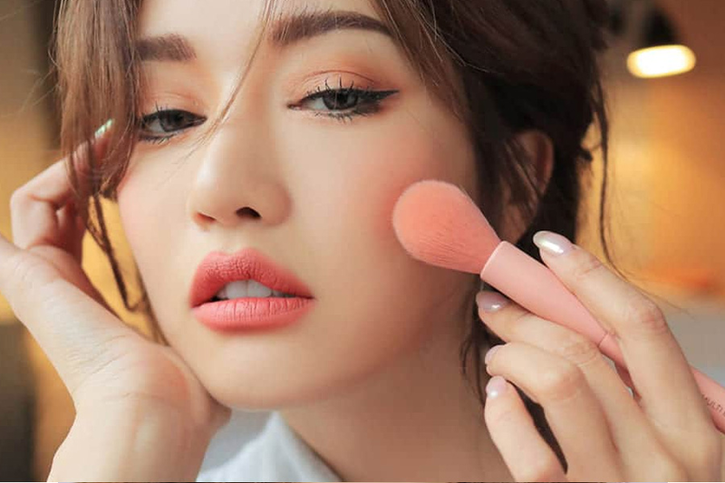 Người phụ nữ trang điểm, dùng cọ phấn má hồng, mắt kẻ eyeliner, môi màu hồng, trong không gian ấm áp.