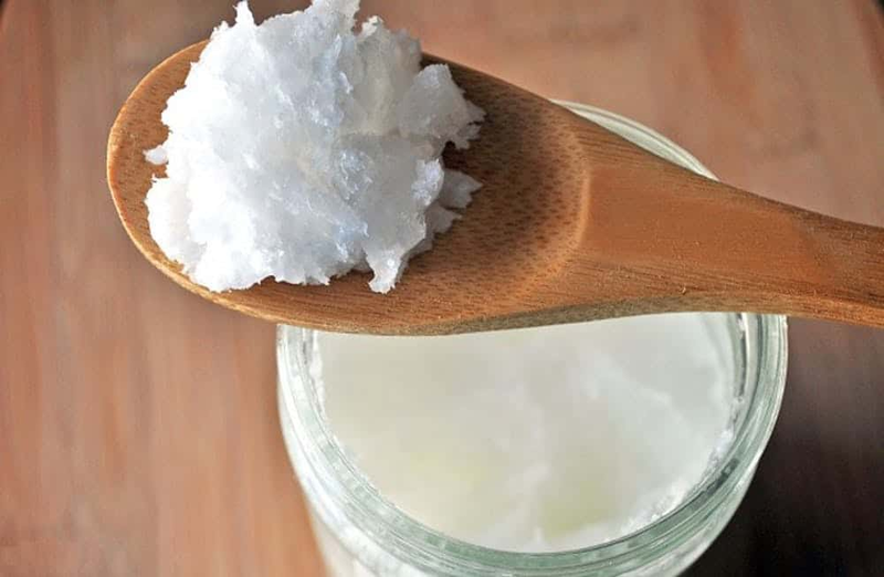 Một thìa gỗ chứa chất trắng mịn, đặt trên miệng lọ thủy tinh đựng cùng loại chất, trên nền gỗ.