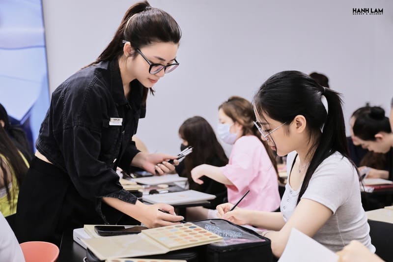Người phụ nữ hướng dẫn học viên trang điểm trong lớp học, xung quanh có nhiều người tham gia.
