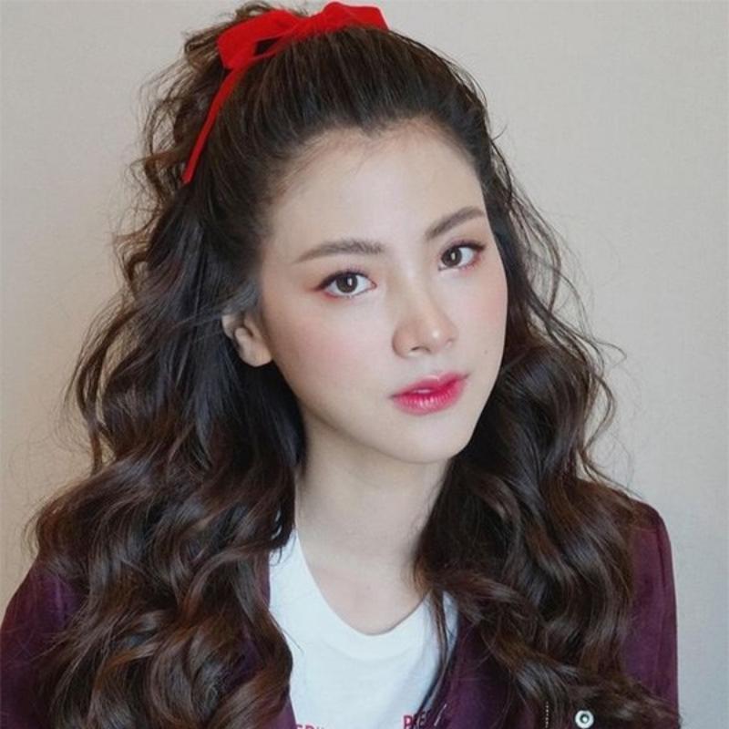 Một người với mái tóc xoăn dài, buộc nơ đỏ, mặc áo trắng và áo khoác tím, nhìn thẳng vào máy ảnh.