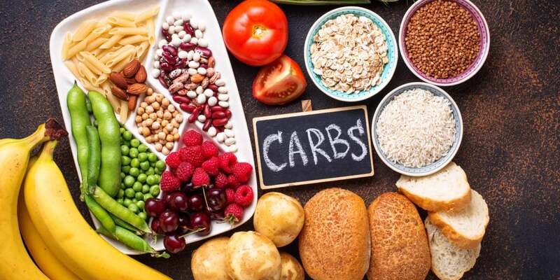 Các loại thực phẩm giàu carbohydrate như bánh mì, trái cây, ngũ cốc và rau củ được bày trên bàn.