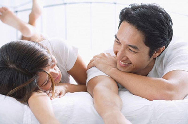 nam-nu-dung-cung-dau-goi Hai người nằm trên giường, mặc áo trắng, cười đùa vui vẻ trong không gian sáng sủa.