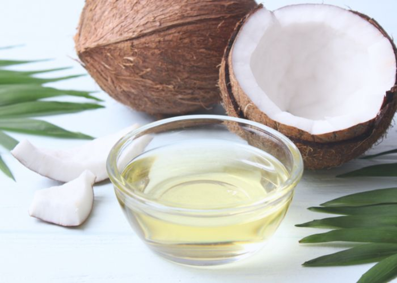 Bát dầu dừa đặt cạnh nửa quả dừa và các lát dừa, trên nền trắng với vài lá xanh.