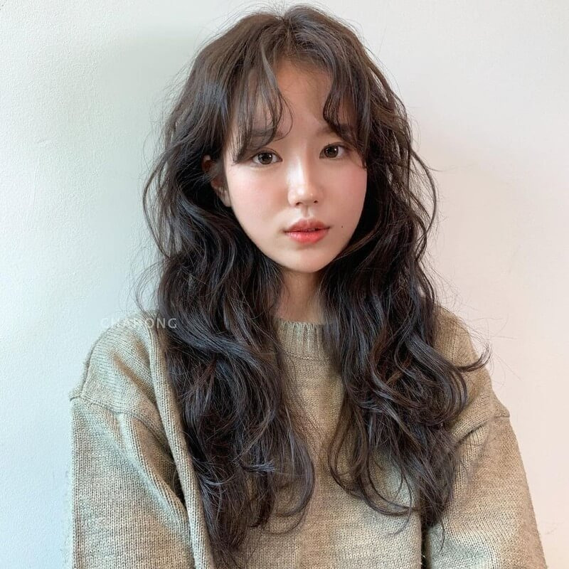 Người phụ nữ trẻ với mái tóc dài uốn sóng nhẹ, mặc áo len màu be, đứng trước nền tường trắng.