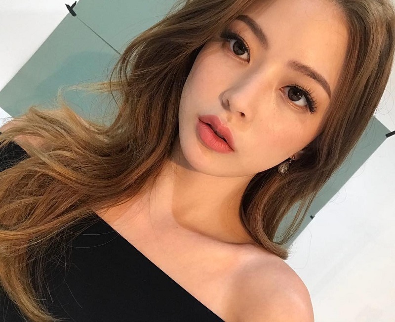 Người phụ nữ tóc dài, mặc áo trễ vai màu đen, đứng trước nền trắng và xanh nhạt.