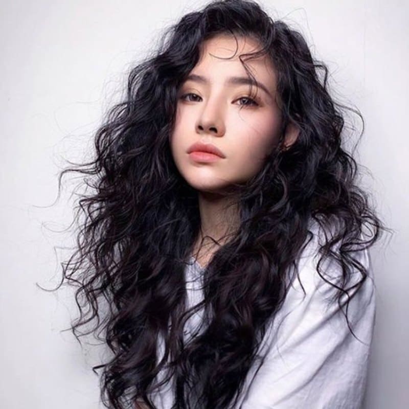 Người phụ nữ với mái tóc xoăn dài, mặc áo trắng, nhìn thẳng về phía trước với biểu cảm nghiêm túc.