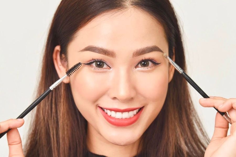 Người phụ nữ cười, cầm cọ trang điểm gần mắt và lông mày, nền trắng.
