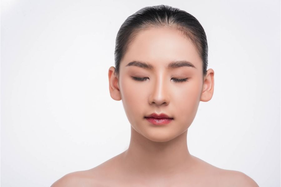 Người phụ nữ trẻ với mắt nhắm, tóc búi gọn, nền trắng đơn giản.