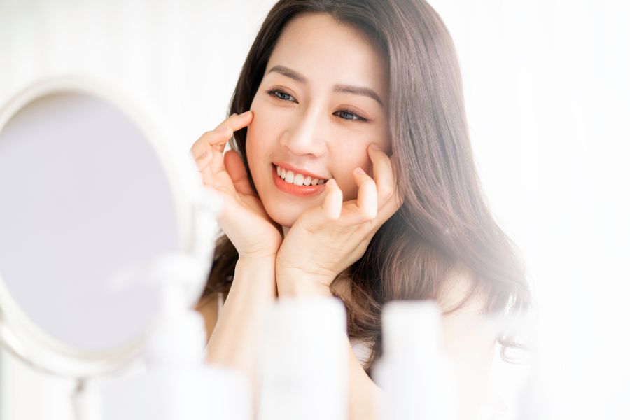 Người phụ nữ cười nhìn vào gương, xung quanh là các chai mỹ phẩm, trong ánh sáng tự nhiên.