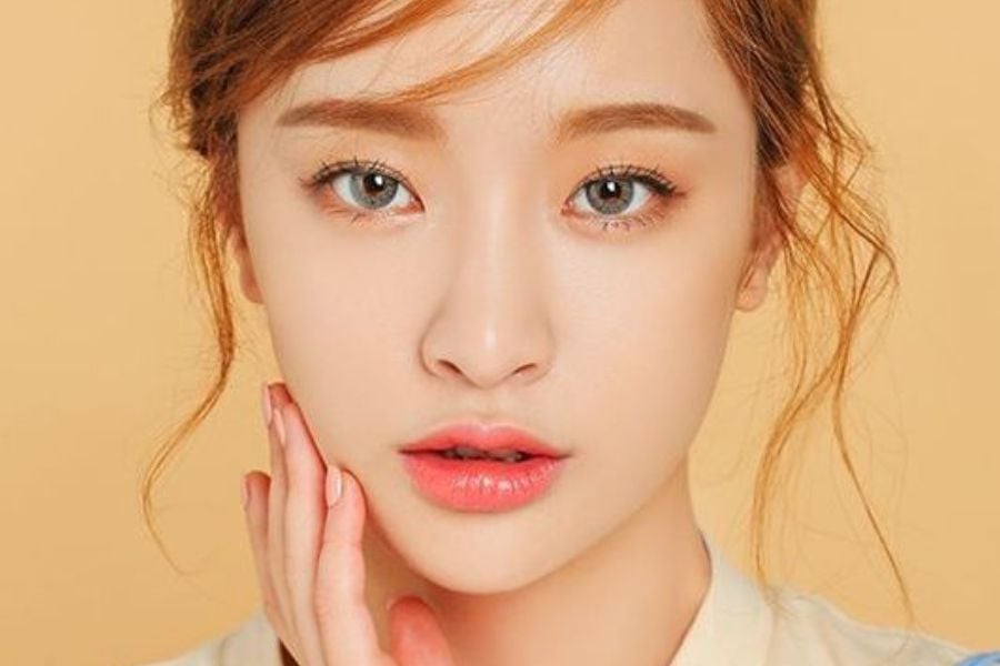 Người phụ nữ trang điểm nhẹ nhàng, tóc buộc hờ, tay chạm nhẹ lên má, nền màu cam nhạt.