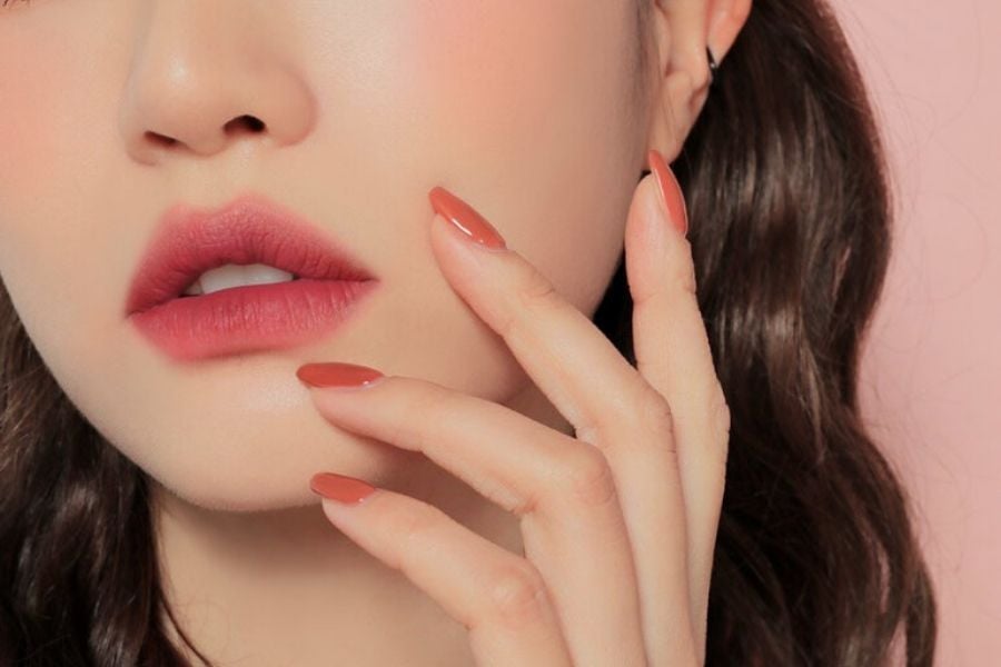 Cận cảnh môi và tay người với móng tay sơn màu đỏ, nền hồng nhạt.
