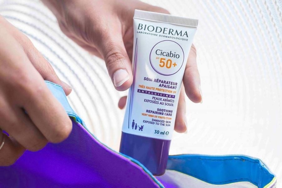 Người cầm tuýp kem chống nắng Bioderma Cicabio SPF 50+ bên cạnh túi màu xanh.