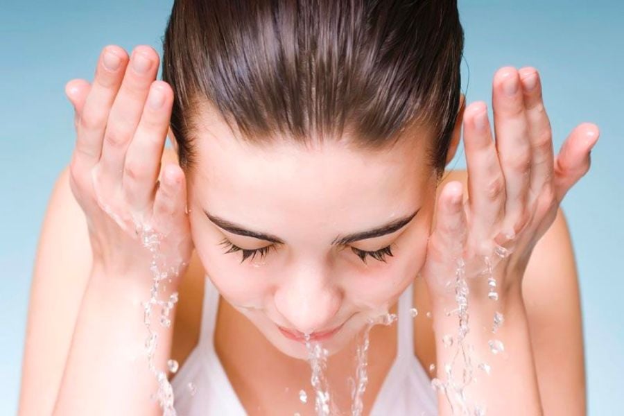 Người đang rửa mặt bằng nước, hai tay nâng nước lên gần mặt, nền xanh nhạt.