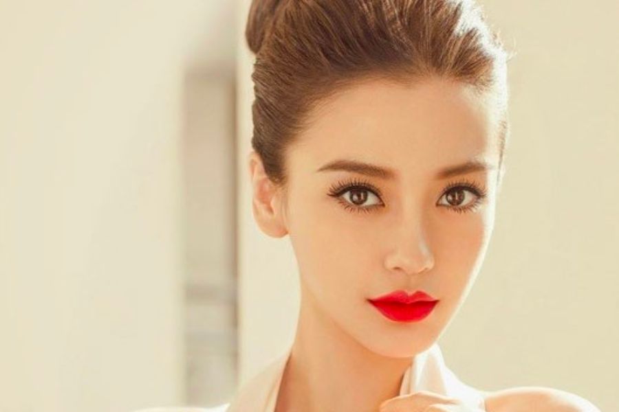 Một người phụ nữ với mái tóc búi cao, trang điểm đậm với son môi đỏ, nhìn thẳng vào ống kính trong nền sáng.