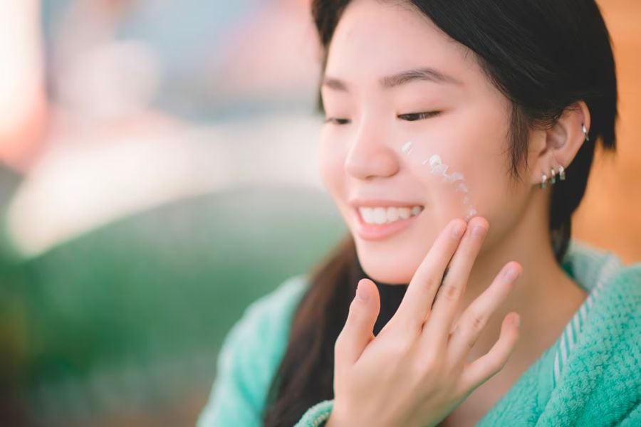 Người phụ nữ đang thoa kem dưỡng lên má, mặc áo xanh, cười tươi trong không gian sáng.
