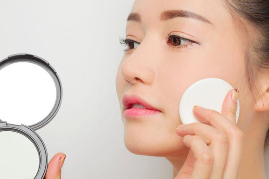 Người phụ nữ đang dùng bông phấn trang điểm lên mặt, tay cầm hộp phấn mở nắp.