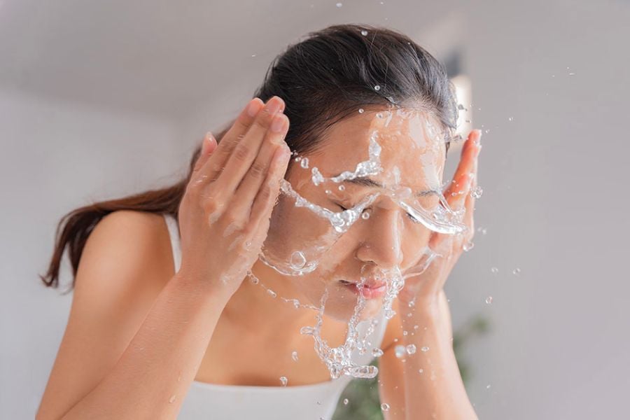 Người đang rửa mặt với nước, tay áp lên hai bên mặt, nước bắn tung tóe xung quanh.