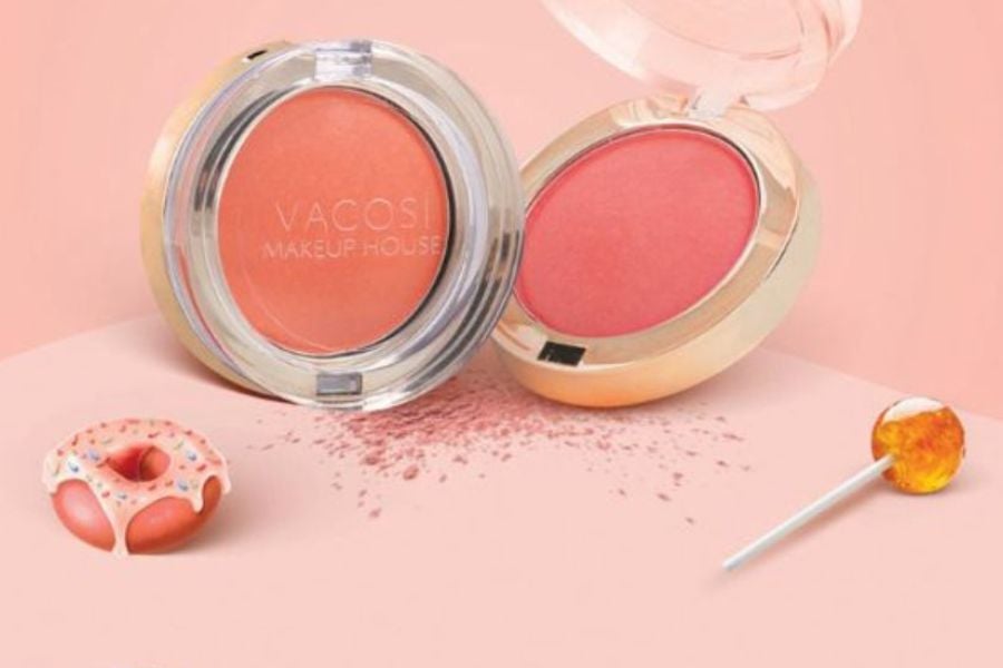 Hộp phấn trang điểm màu cam hồng trên nền màu hồng nhạt, cạnh một chiếc bánh donut và kẹo mút.