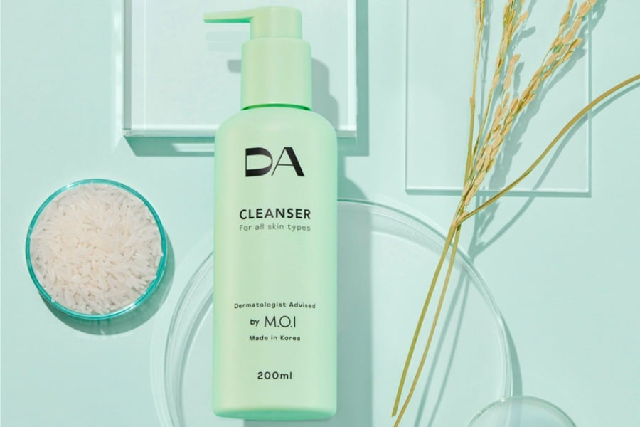 Chai sữa rửa mặt màu xanh lá cây đặt cạnh bát gạo và nhánh lúa trên nền xanh pastel.