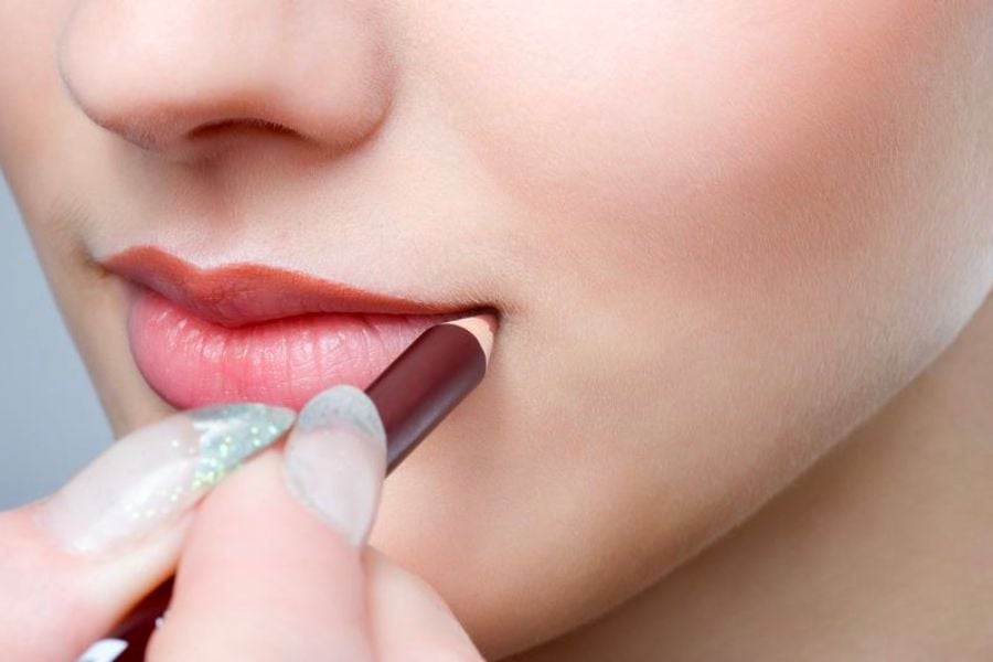 Người đang dùng bút kẻ môi màu hồng nhạt để tô viền môi, tạo đường nét rõ ràng cho đôi môi.