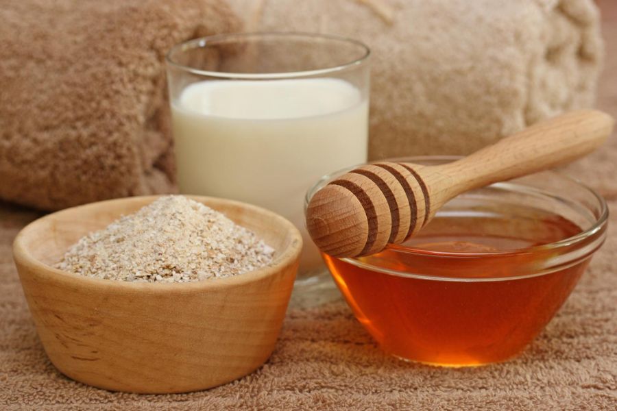 Bột yến mạch, ly sữa và chén mật ong cùng dụng cụ gỗ trên nền khăn tắm màu nâu.