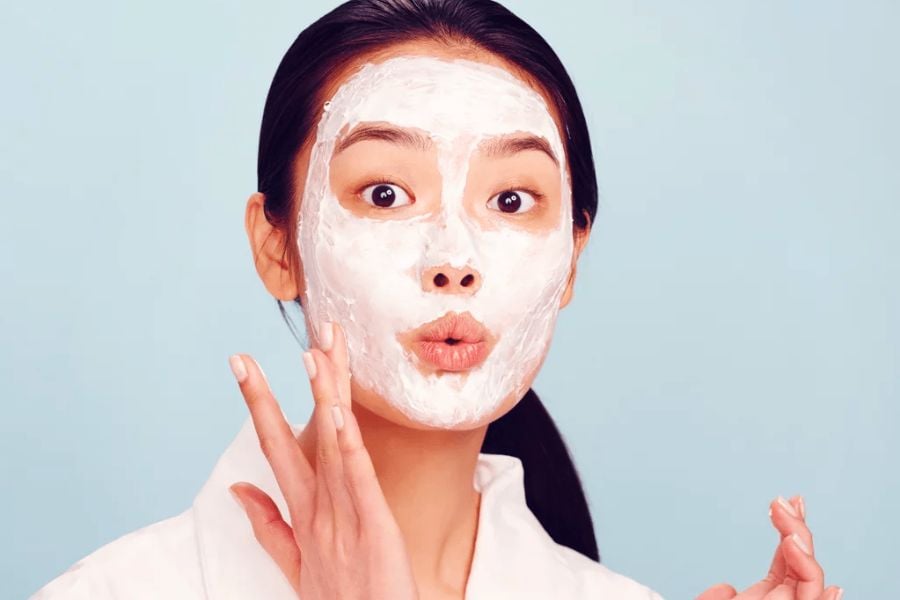 Người đang thoa mặt nạ dưỡng da màu trắng, biểu cảm khuôn mặt ngạc nhiên, nền xanh nhạt.