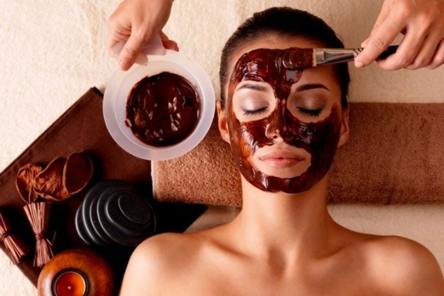 Người đang được đắp mặt nạ sô cô la trong một liệu trình chăm sóc da, tay cầm cọ và bát hỗn hợp.