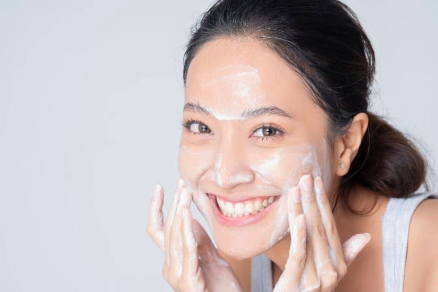 Người phụ nữ đang rửa mặt với sữa rửa mặt, tay tạo bọt trên da, cười tươi, nền trắng.