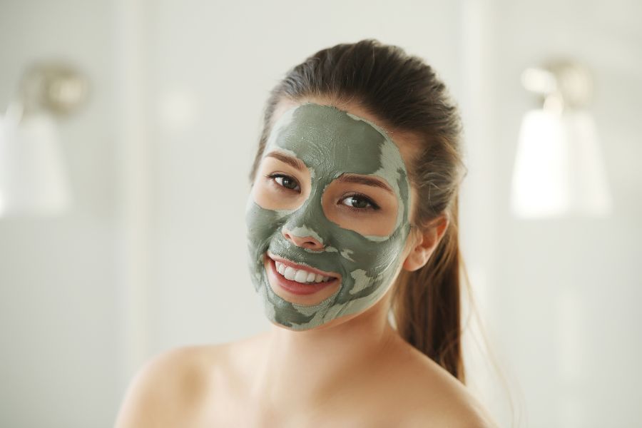 Người đang đắp mặt nạ dưỡng da màu xanh, mỉm cười, nền mờ với ánh sáng dịu.