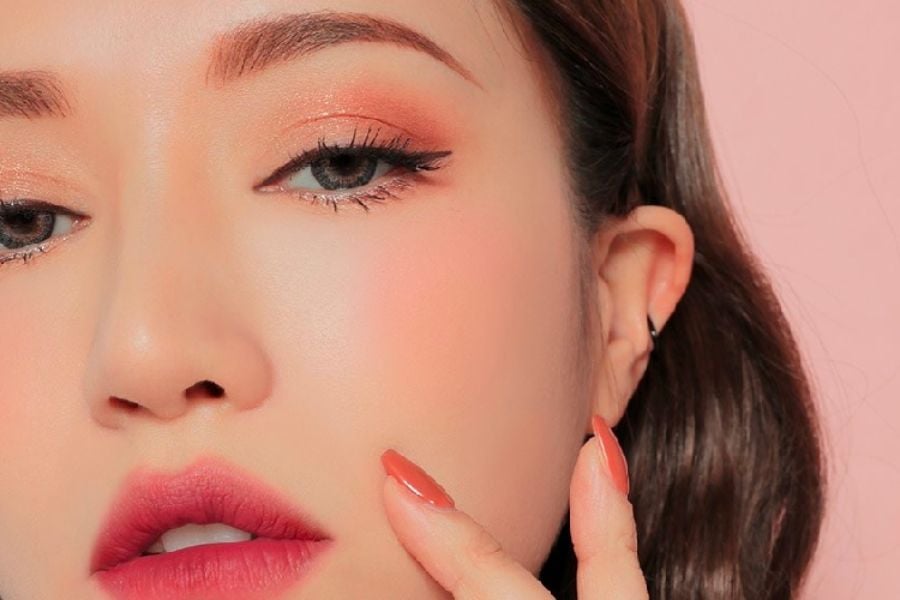 Cận cảnh khuôn mặt với trang điểm mắt và môi nổi bật, tay chạm nhẹ lên má, nền màu hồng nhạt.