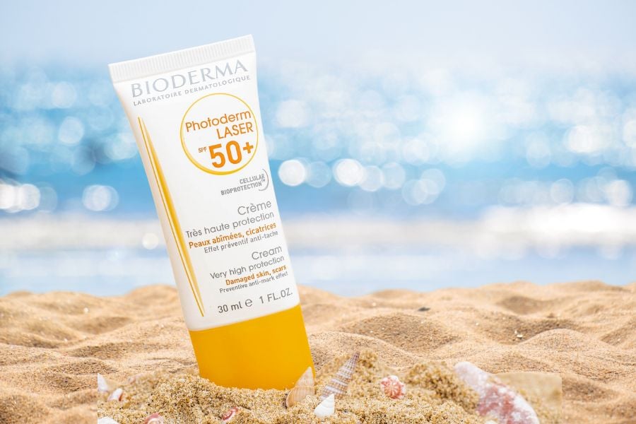 Tuýp kem chống nắng SPF 50+ trên bãi cát, ánh sáng phản chiếu tạo hiệu ứng lung linh từ nước biển phía sau.