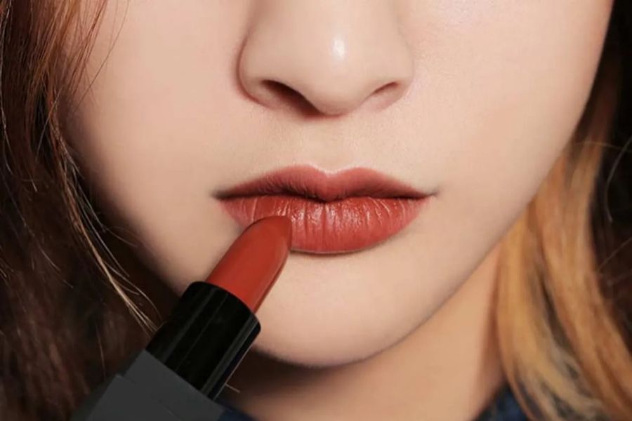 Cận cảnh một người đang thoa son môi màu đỏ cam lên môi.