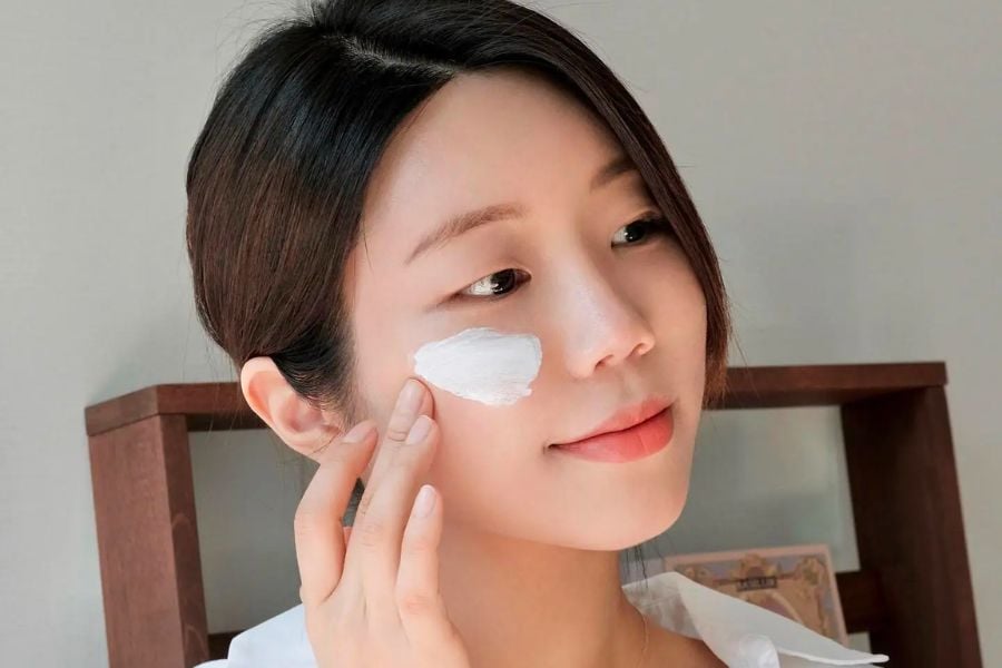 Người phụ nữ thoa kem lên má, tóc buộc gọn, mặc áo trắng, đứng trước kệ gỗ.