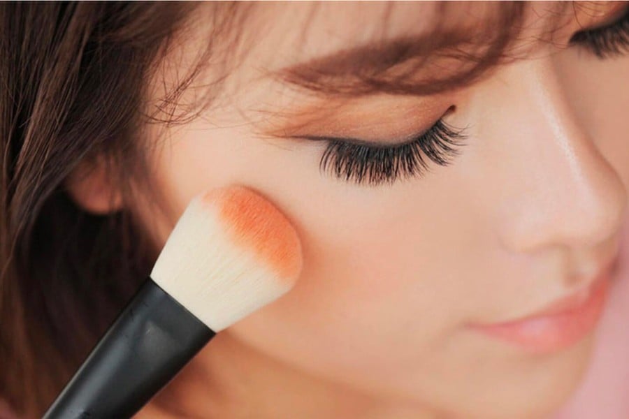 Cận cảnh một người đang dùng cọ trang điểm để thoa phấn má hồng lên má.