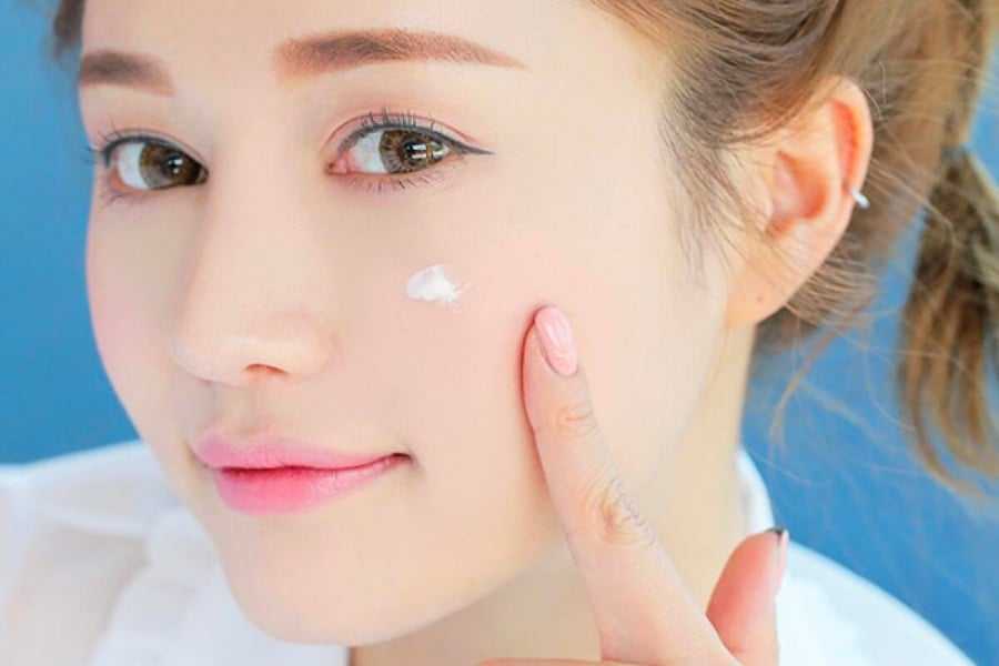 Người đang thoa kem dưỡng da lên má, nền xanh, tóc buộc nhẹ, mặc áo trắng.