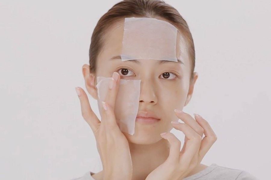 Người đang đắp mặt nạ giấy lên trán và má, tay chạm vào mặt nạ, nền trắng.