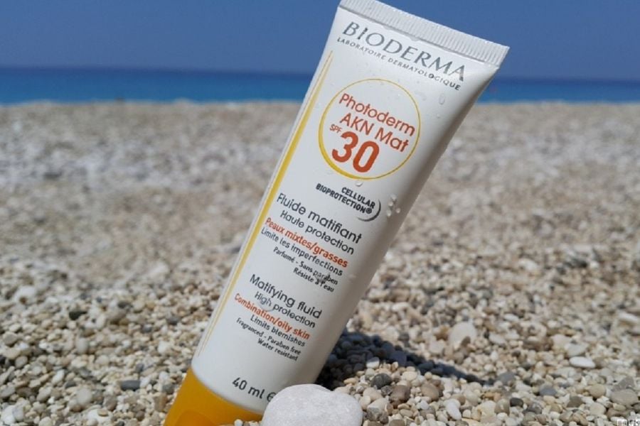 Tuýp kem chống nắng SPF 30 nằm trên bãi biển sỏi, nền biển xanh và bầu trời trong.