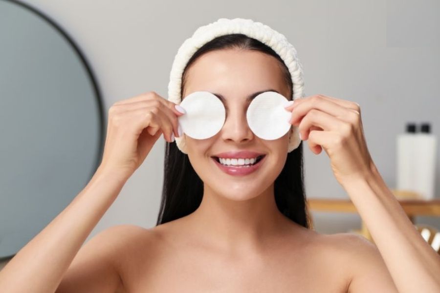 Người phụ nữ cười, dùng bông tẩy trang che mắt, đội băng đô trắng trong phòng tắm.