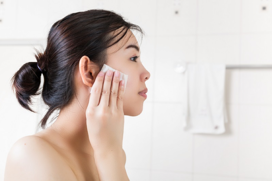 Người phụ nữ dùng bông tẩy trang trên mặt trong phòng tắm với nền tường trắng.