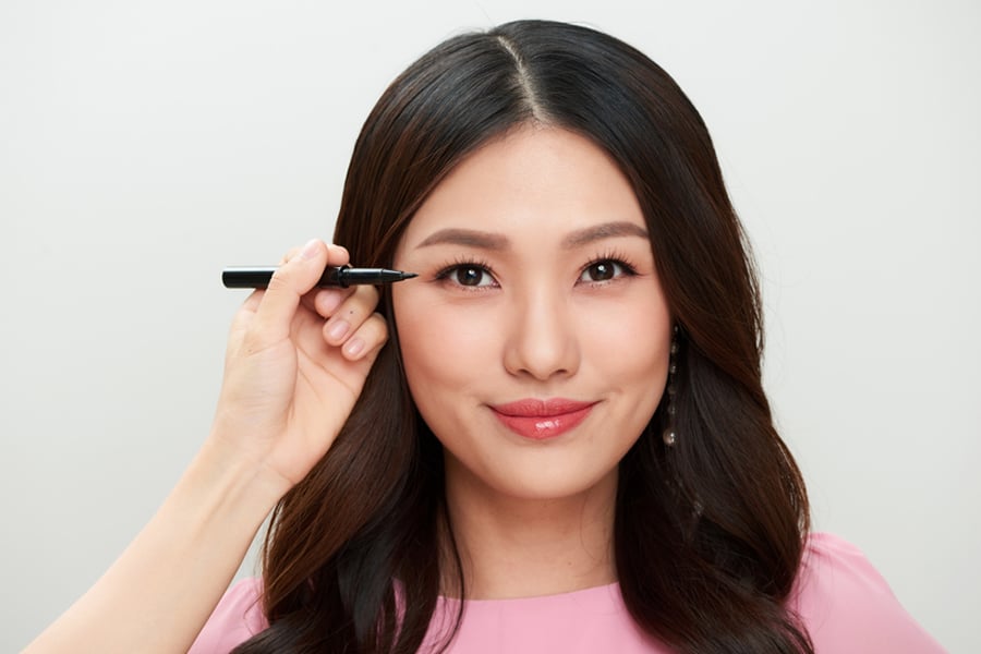 Người phụ nữ đang được trang điểm mắt bằng bút kẻ mắt, với biểu cảm mỉm cười, mặc áo màu hồng.