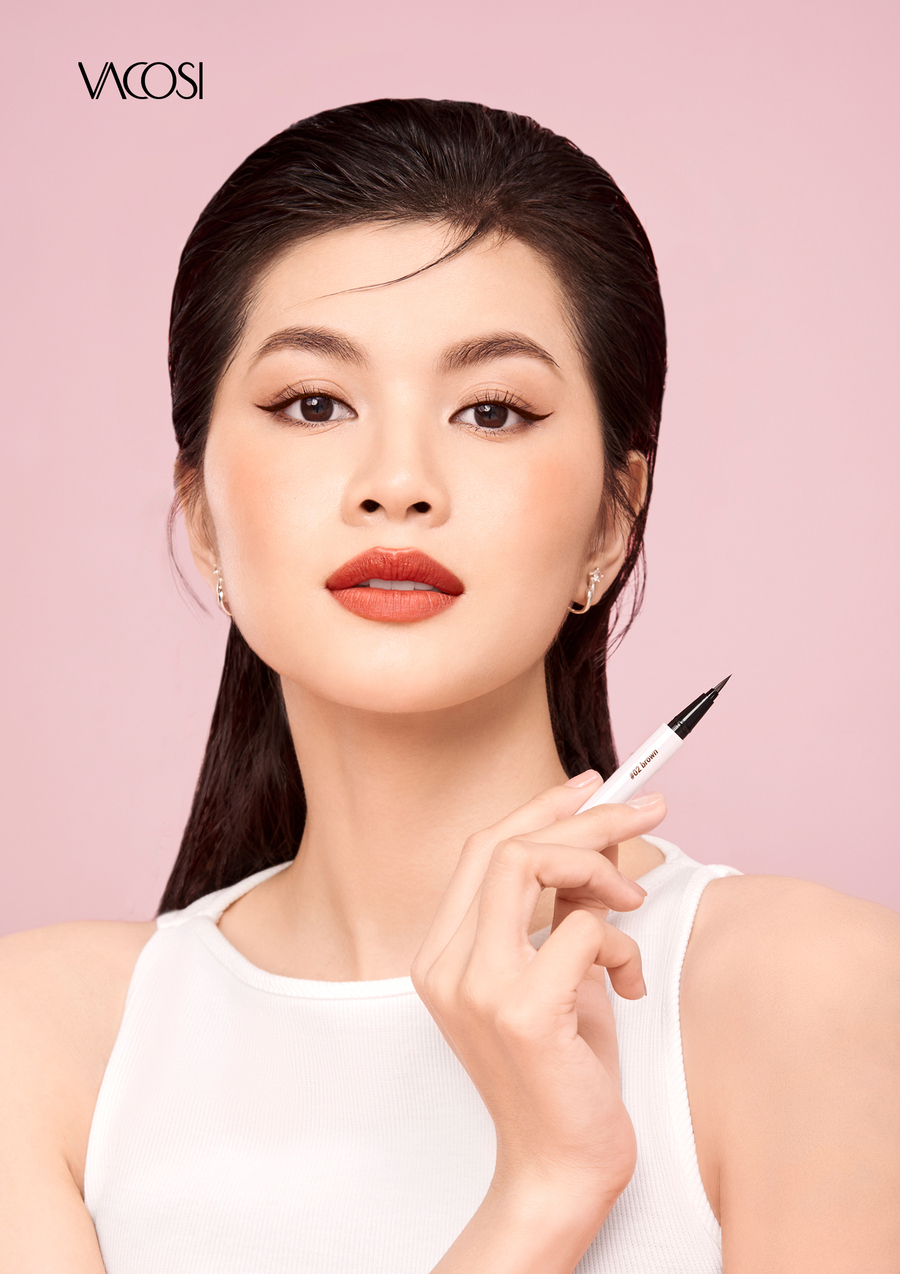 Người phụ nữ cầm bút kẻ mắt, trang điểm tinh tế, nền hồng nhạt, mặc áo trắng không tay.