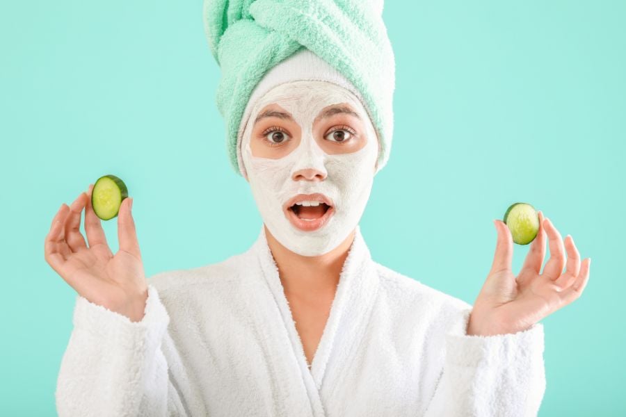 Người đắp mặt nạ đất sét, quấn khăn xanh, cầm hai lát dưa chuột, mặc áo choàng trắng, nền xanh.