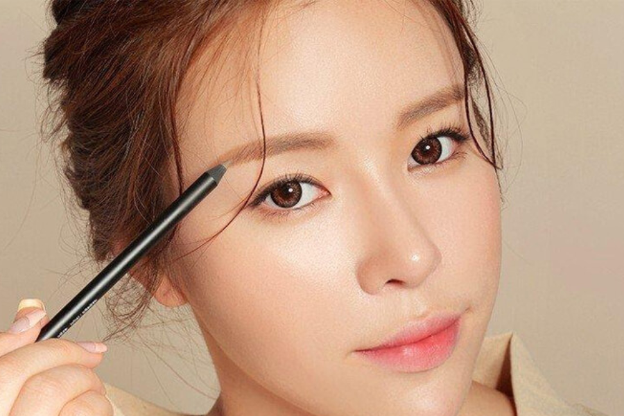 Người phụ nữ đang kẻ lông mày bằng bút chì trang điểm, nền da sáng và tóc buộc gọn.