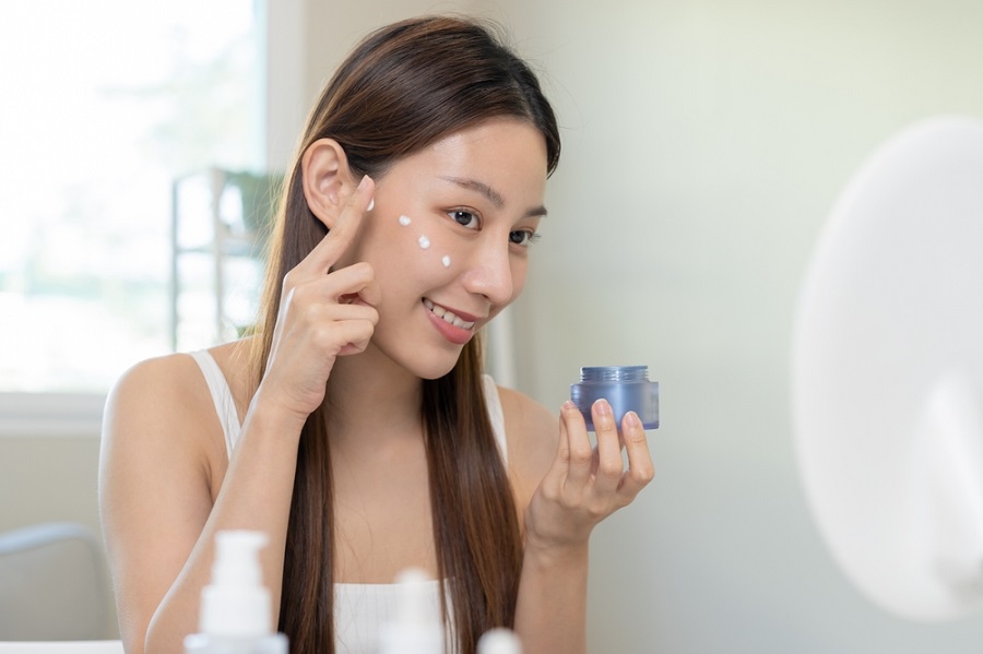 Người phụ nữ đang thoa kem dưỡng da lên mặt, cầm hũ kem và mỉm cười trước gương trong phòng sáng.