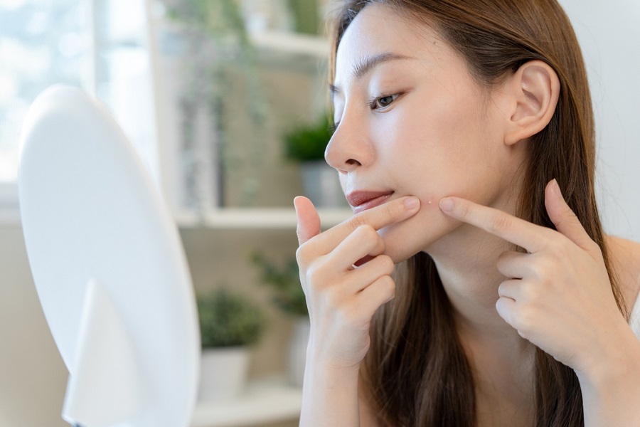 Người phụ nữ nhìn vào gương, dùng hai ngón tay chạm vào da mặt, có thể đang xử lý mụn.