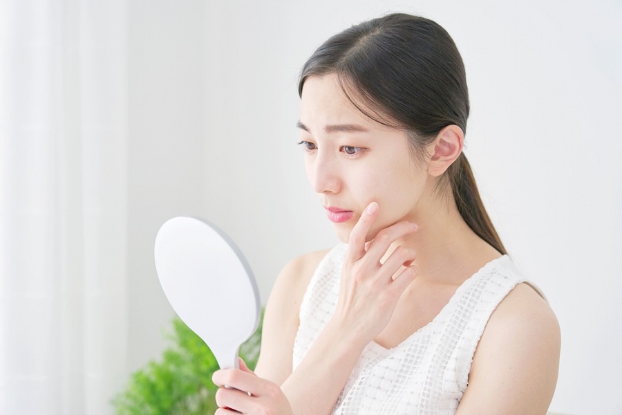 Người phụ nữ đang cầm gương soi, chạm nhẹ vào mặt, biểu cảm suy tư trong không gian sáng sủa.