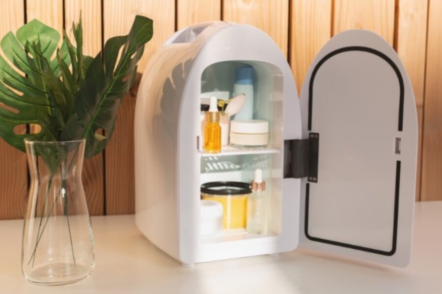 Tủ lạnh mini mở cửa chứa mỹ phẩm, cạnh đó là bình hoa lá xanh, nền gỗ sáng màu.
