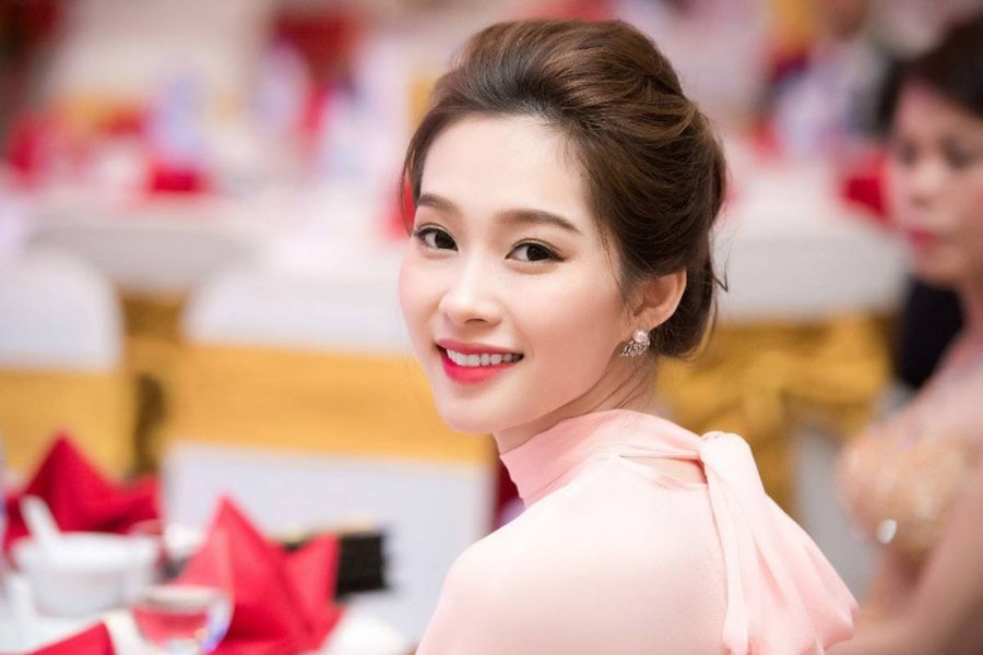 Người phụ nữ mặc áo dài hồng, mỉm cười, ngồi trong không gian sang trọng với khăn trải bàn đỏ và vàng.
