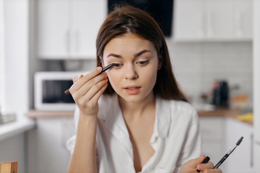 Người phụ nữ trong bếp đang trang điểm mắt bằng bút kẻ mắt, mặc áo trắng.
