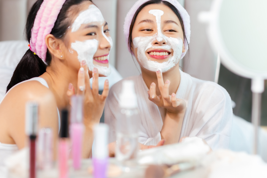 Hai người đang đắp mặt nạ dưỡng da, mỉm cười trước gương, xung quanh có chai lọ mỹ phẩm.